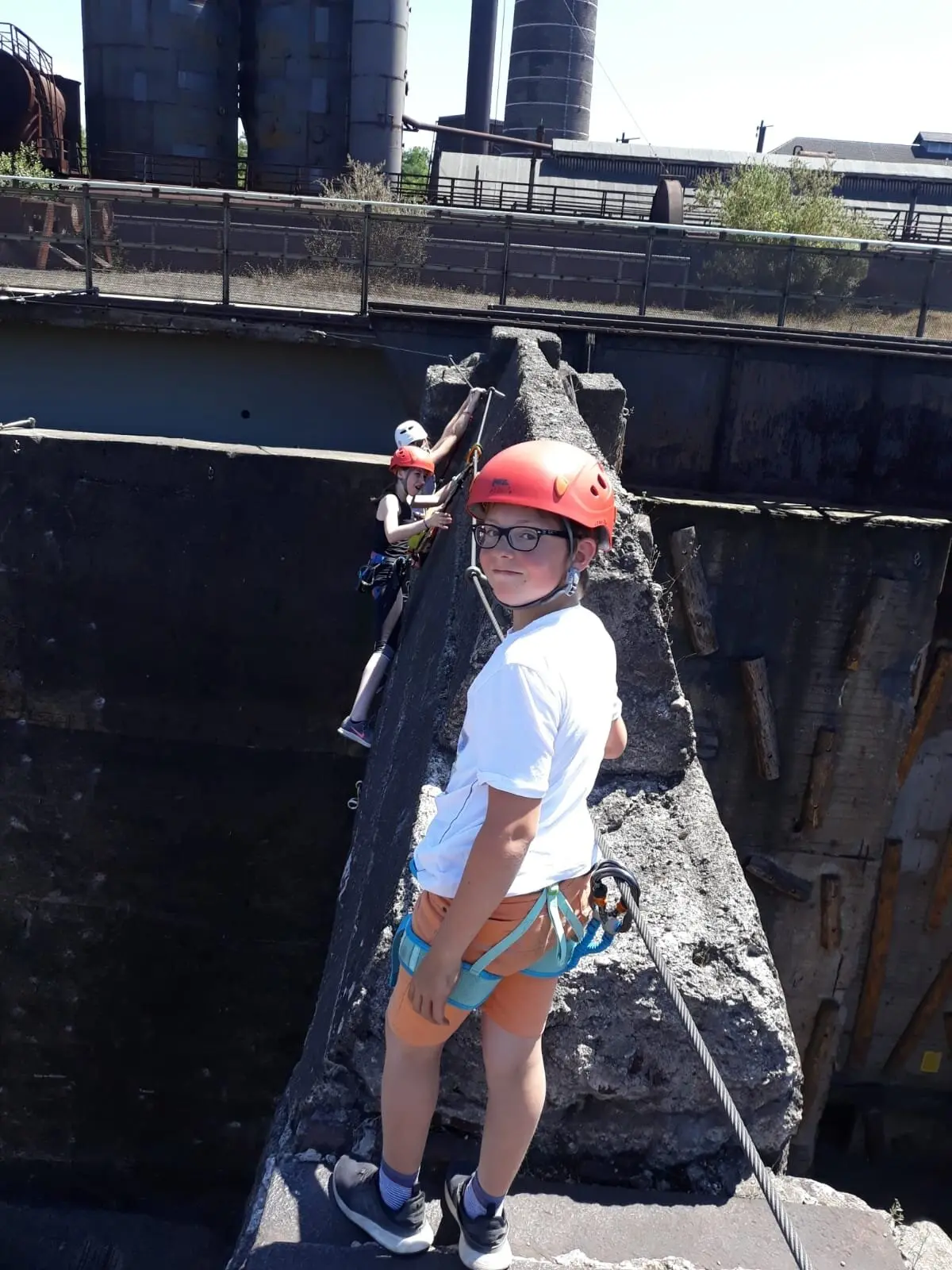 Zwei Kinder mit Helm und Klettergurt stehen gesichert auf einem Betonsteg zwischen alten Industrieanlagen. | © DAV Dortmund