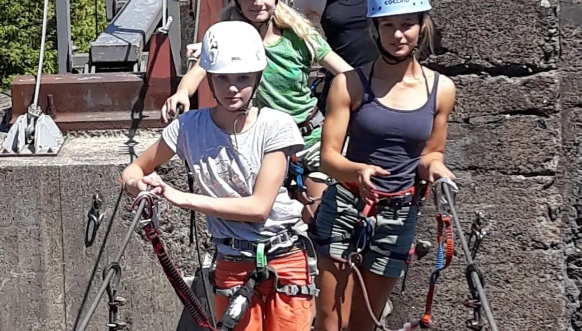 Vier Kinder mit Kletterausrüstung überqueren gesichert eine Seilbrücke an einer Betonwand im Industriegelände. | © DAV Dortmund