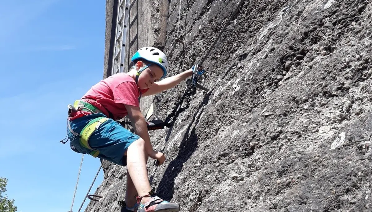 Ein Kind klettert gesichert an einer grauen Outdoor-Kletterwand bei Sonnenschein. | © DAV Dortmund