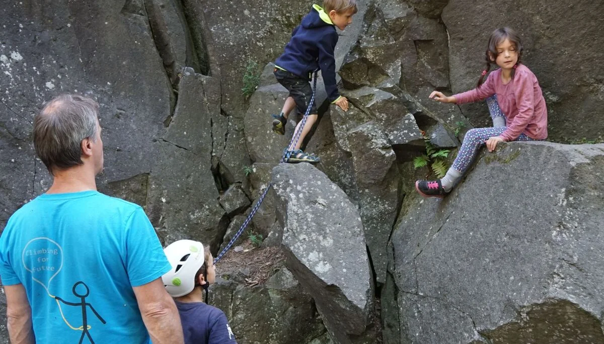 Zwei Kinder klettern zwischen Felsblöcken, ein Erwachsener sichert und beobachtet die Szene. | © DAV Dortmund