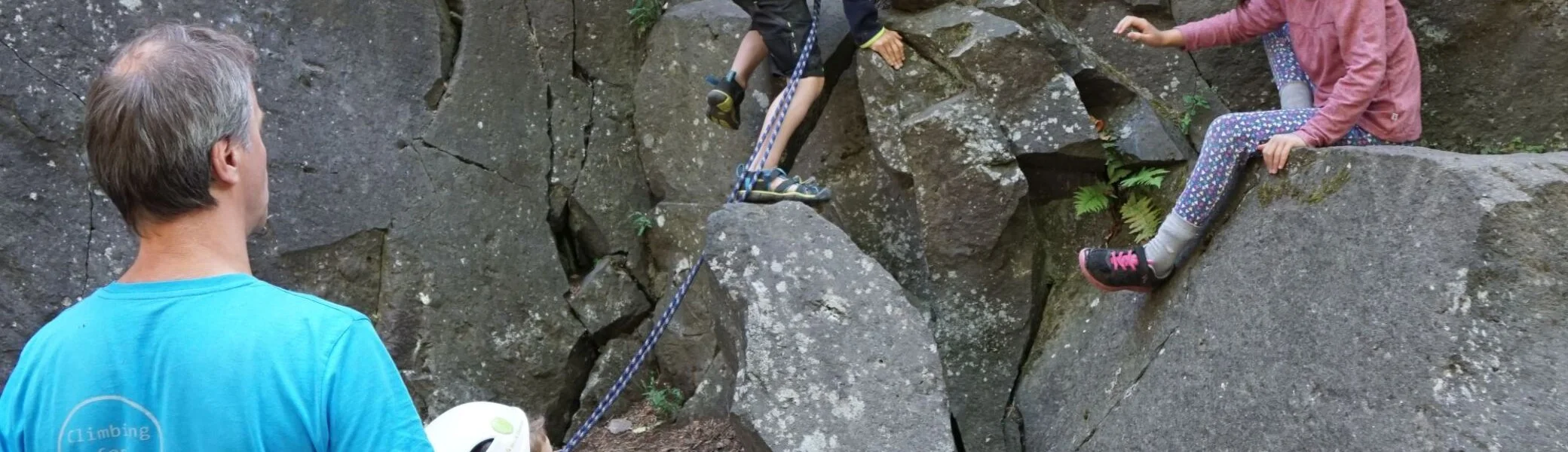 Zwei Kinder klettern zwischen Felsblöcken, ein Erwachsener sichert und beobachtet die Szene. | © DAV Dortmund