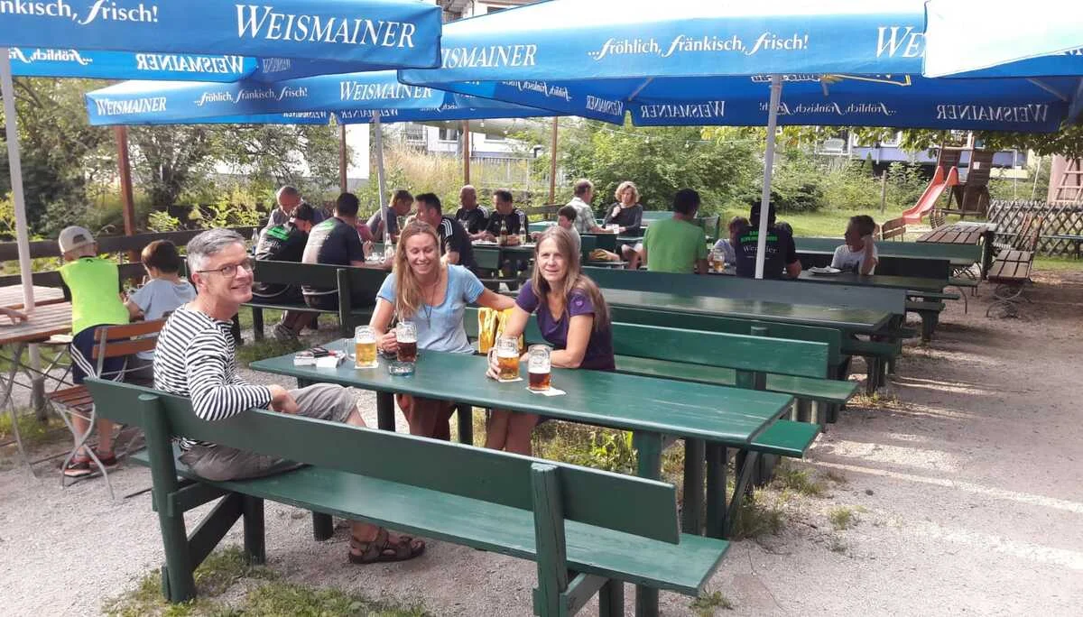 Drei Personen sitzen mit Getränken an einem grünen Tisch im schattigen Biergarten. | © DAV Dortmund