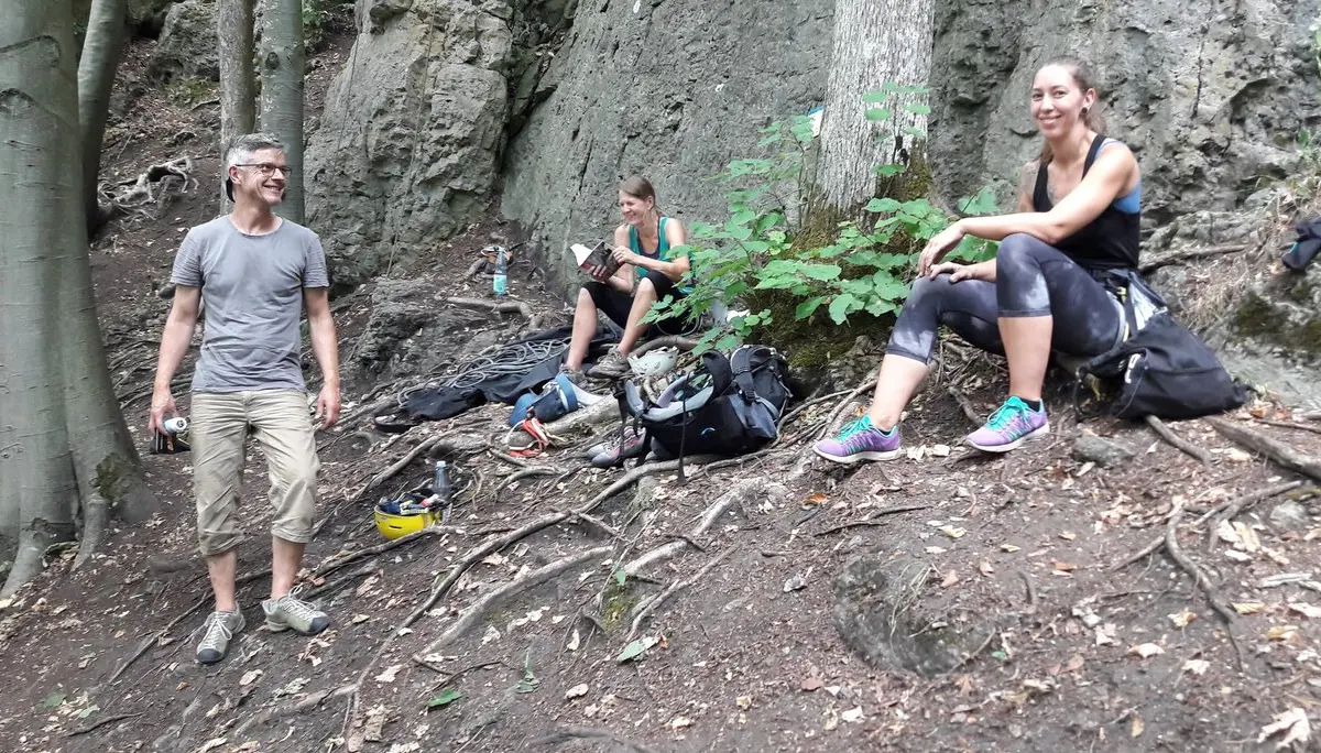Drei Personen sitzen und stehen entspannt im Wald auf dem Waldboden, mit Kletterausrüstung umgeben. | © DAV Dortmund