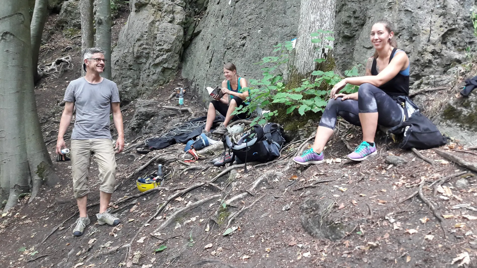 Drei Personen sitzen und stehen entspannt im Wald auf dem Waldboden, mit Kletterausrüstung umgeben. | © DAV Dortmund