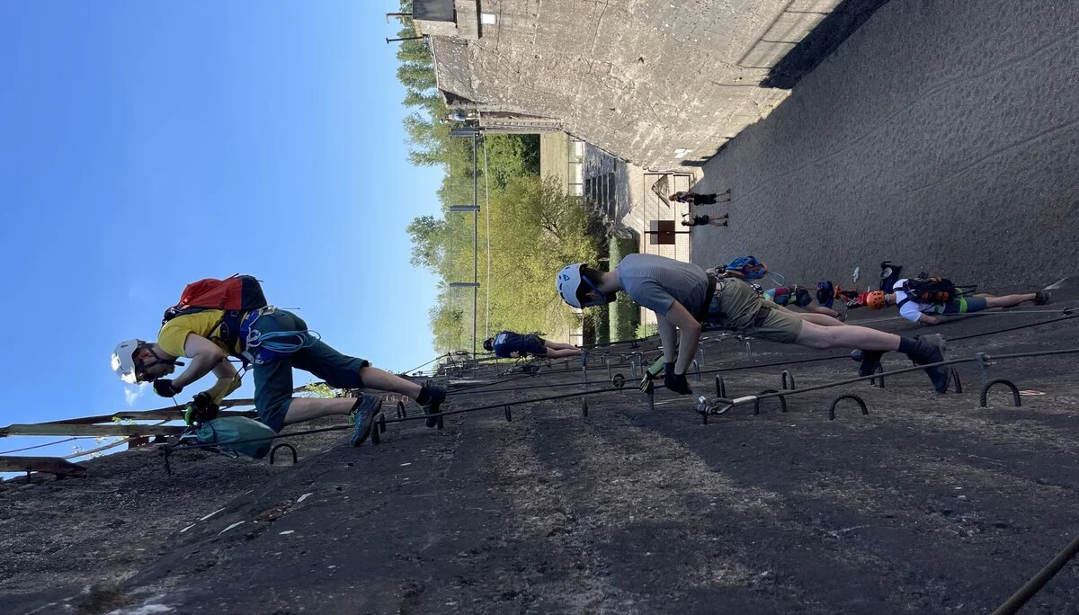 Teilnehmende steigen mit Kletterausrüstung einen künstlichen Klettersteig an einer hohen Betonwand empor. | © DAV Dortmund