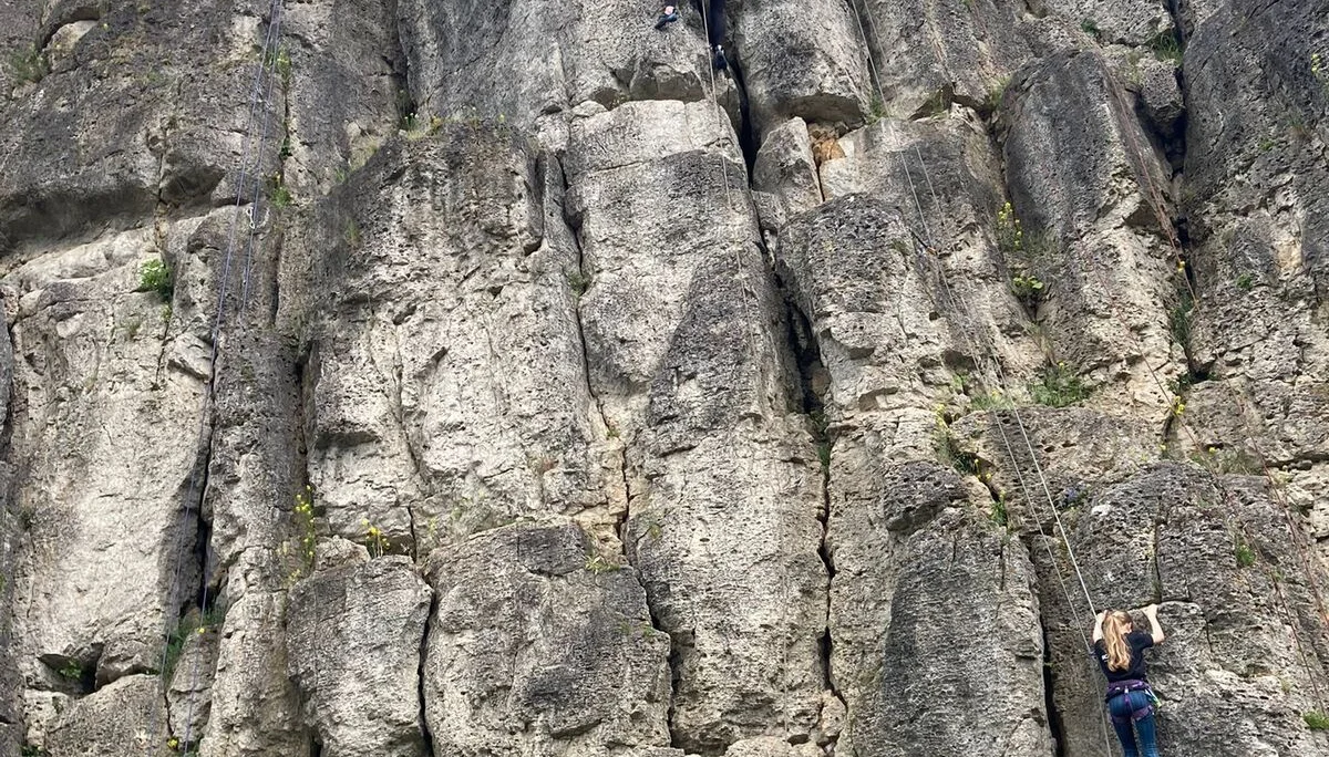 Mehrere Personen klettern an einer hohen, strukturierten Felswand mit Gipfelkreuz oben. | © DAV Dortmund