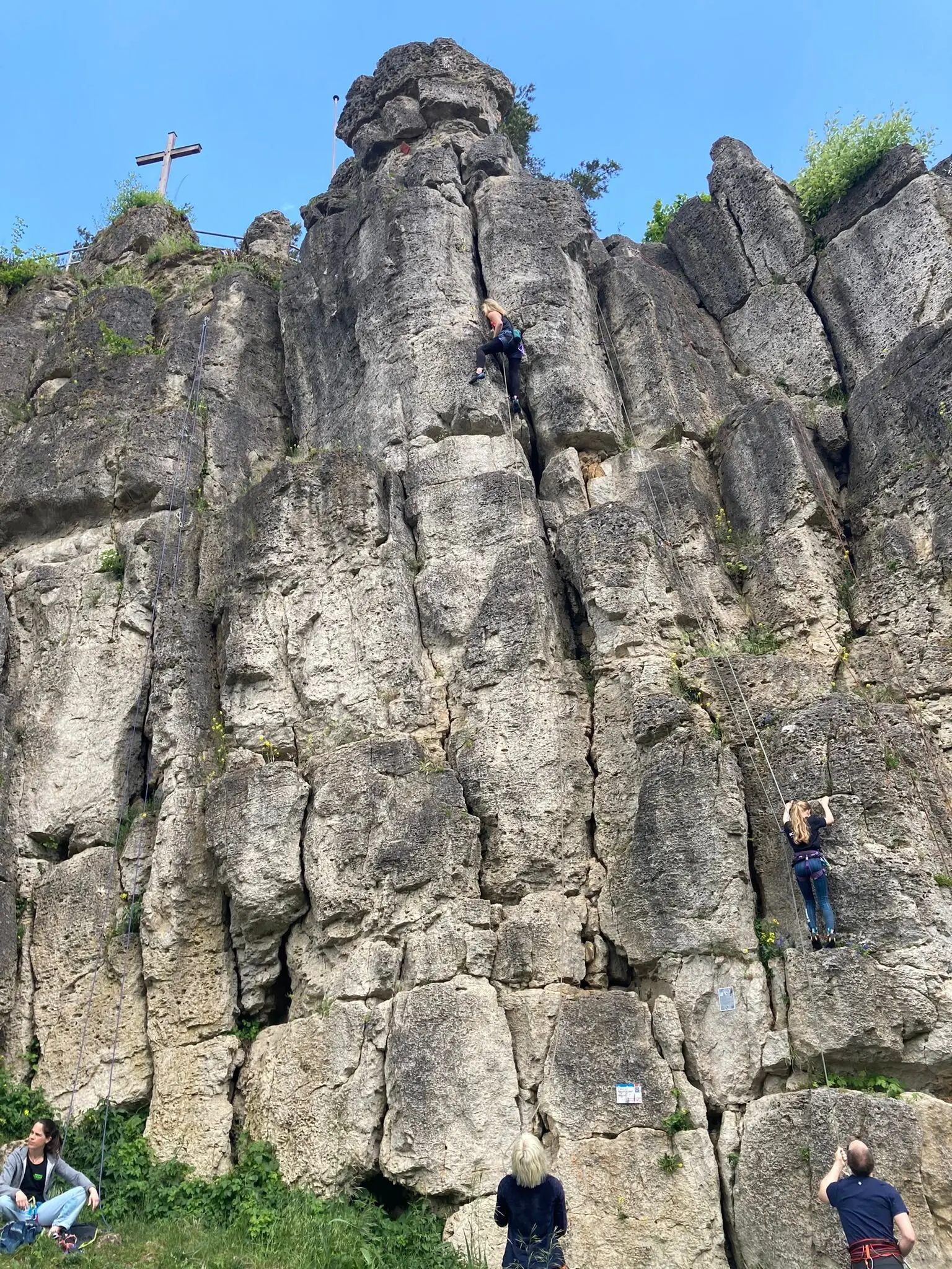 Mehrere Personen klettern an einer hohen, strukturierten Felswand mit Gipfelkreuz oben. | © DAV Dortmund