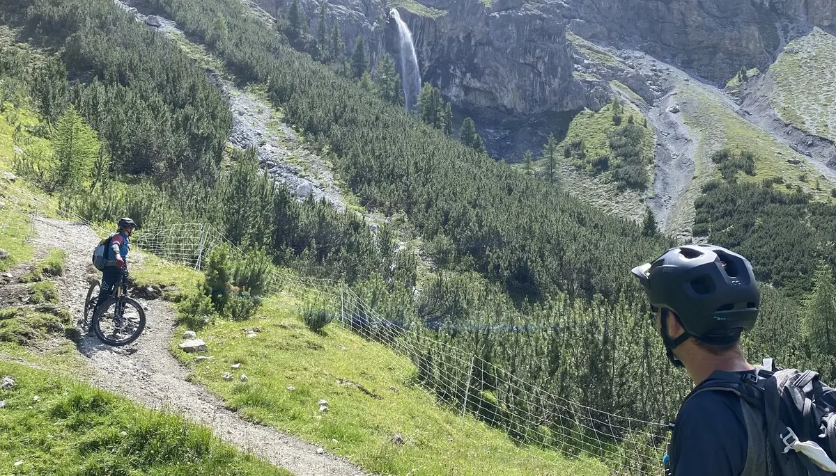 Zwei Mountainbiker auf einem grünen Wanderweg mit Wasserfall und steilen Berghängen im Hintergrund. | © DAV Dortmund