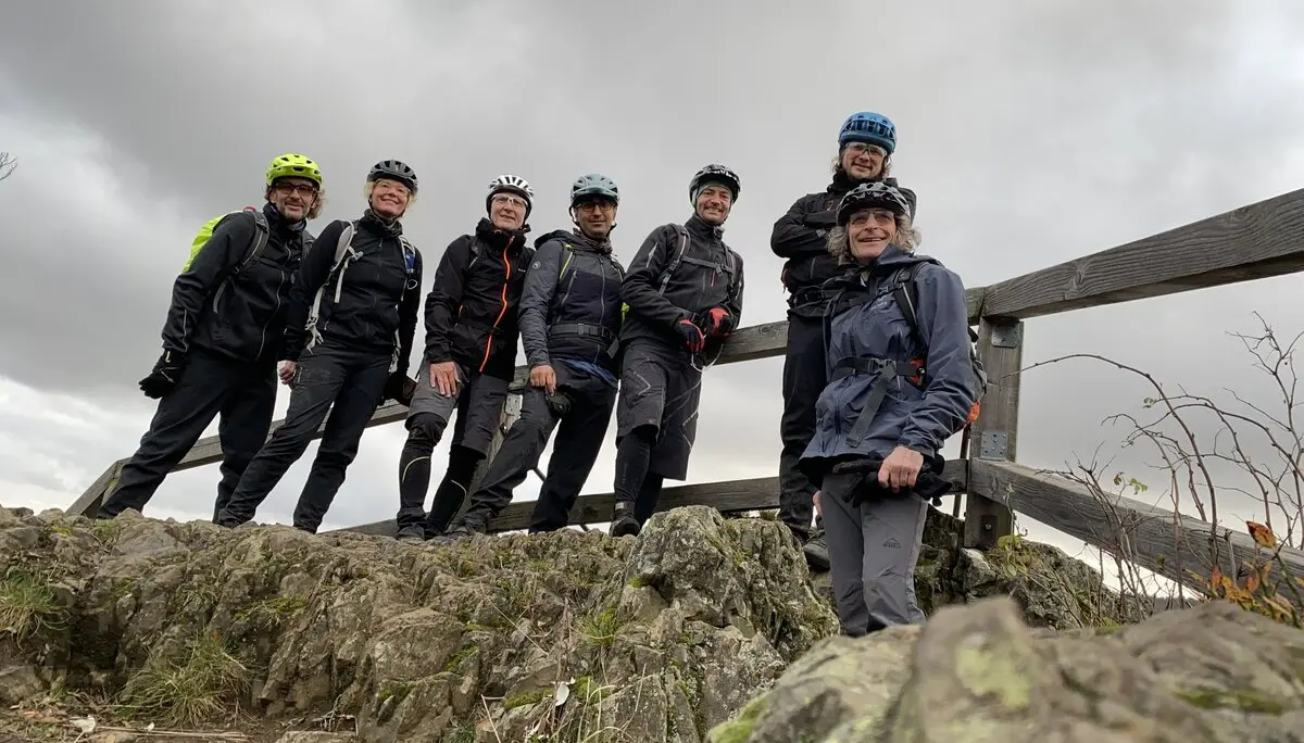 Acht Mountainbiker*innen posieren auf einer felsigen Anhöhe neben einem Holzgeländer bei wolkigem Himmel. | © DAV Dortmund
