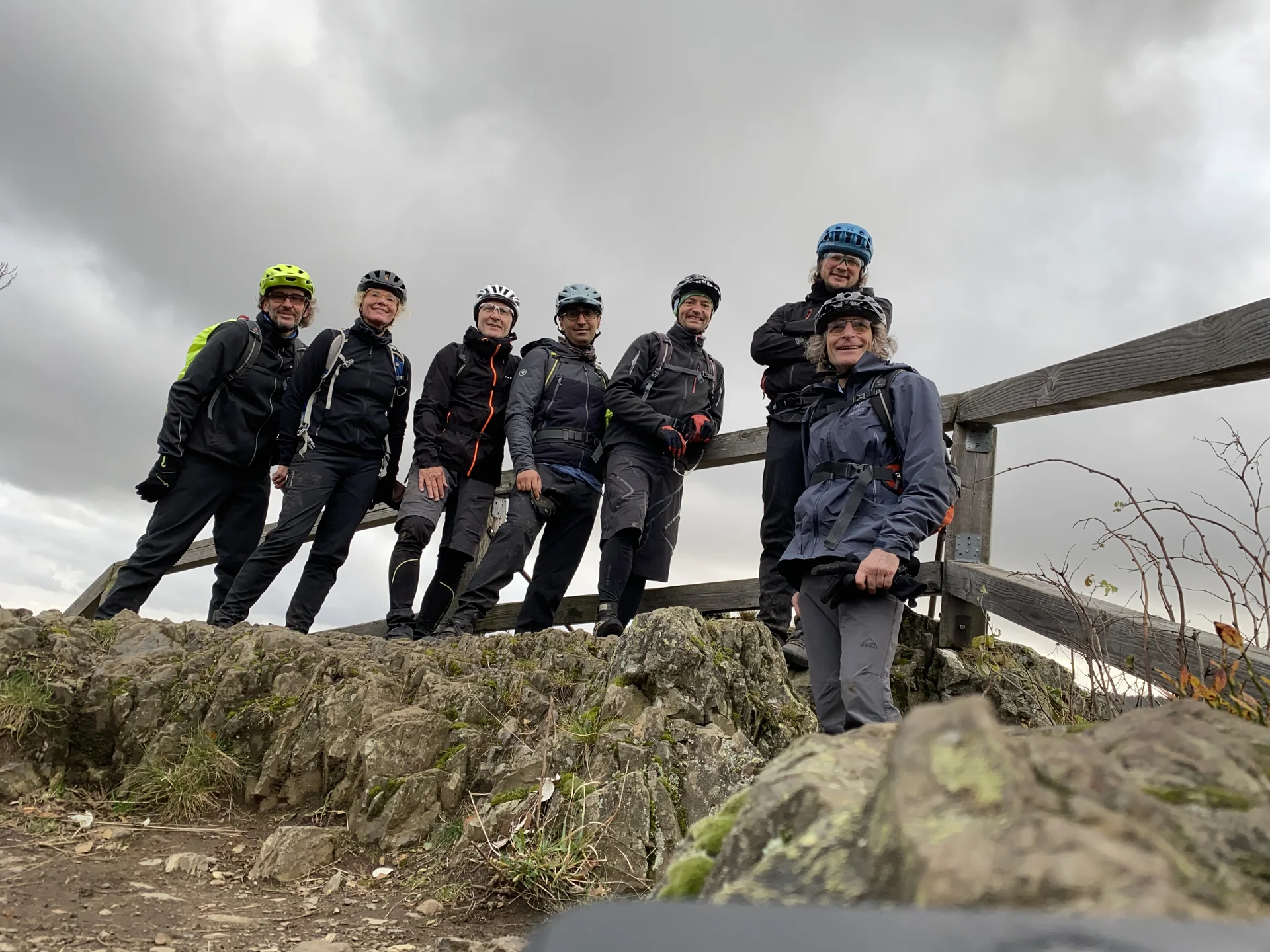 Acht Mountainbiker*innen posieren auf einer felsigen Anhöhe neben einem Holzgeländer bei wolkigem Himmel. | © DAV Dortmund