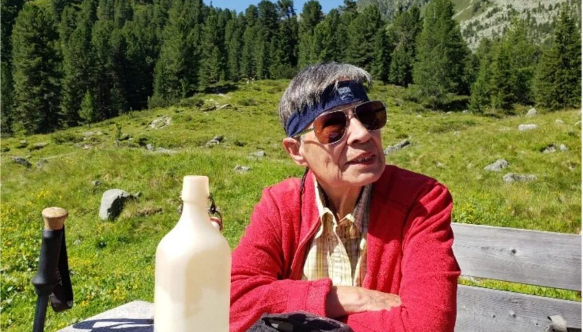 Ältere Frau mit Sonnenbrille und Stirnband sitzt an einem Holztisch auf einer Almwiese. | © DAV Dortmund