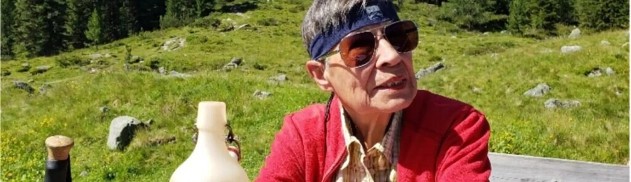 Ältere Frau mit Sonnenbrille und Stirnband sitzt an einem Holztisch auf einer Almwiese. | © DAV Dortmund