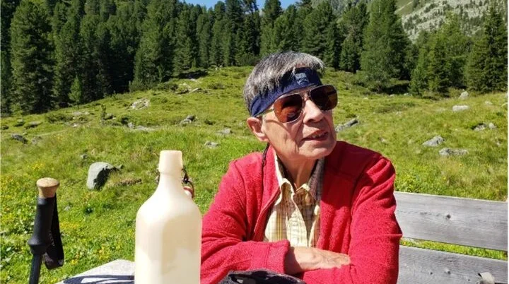 Ältere Frau mit Sonnenbrille und Stirnband sitzt an einem Holztisch auf einer Almwiese. | © DAV Dortmund