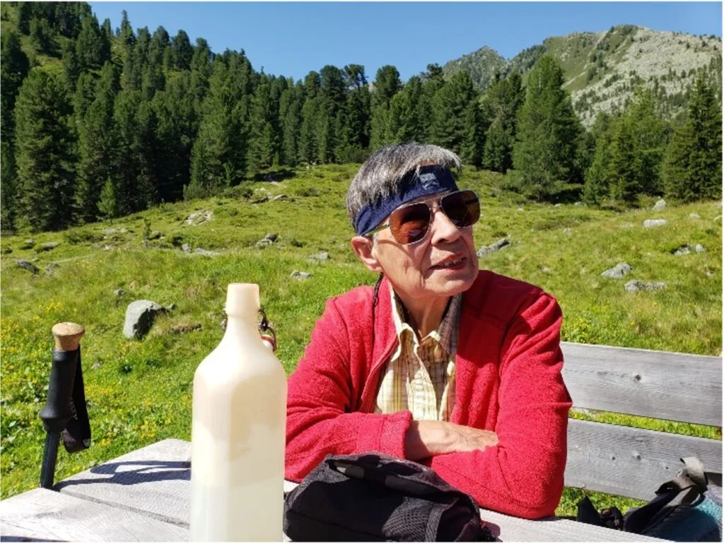 Ältere Frau mit Sonnenbrille und Stirnband sitzt an einem Holztisch auf einer Almwiese. | © DAV Dortmund