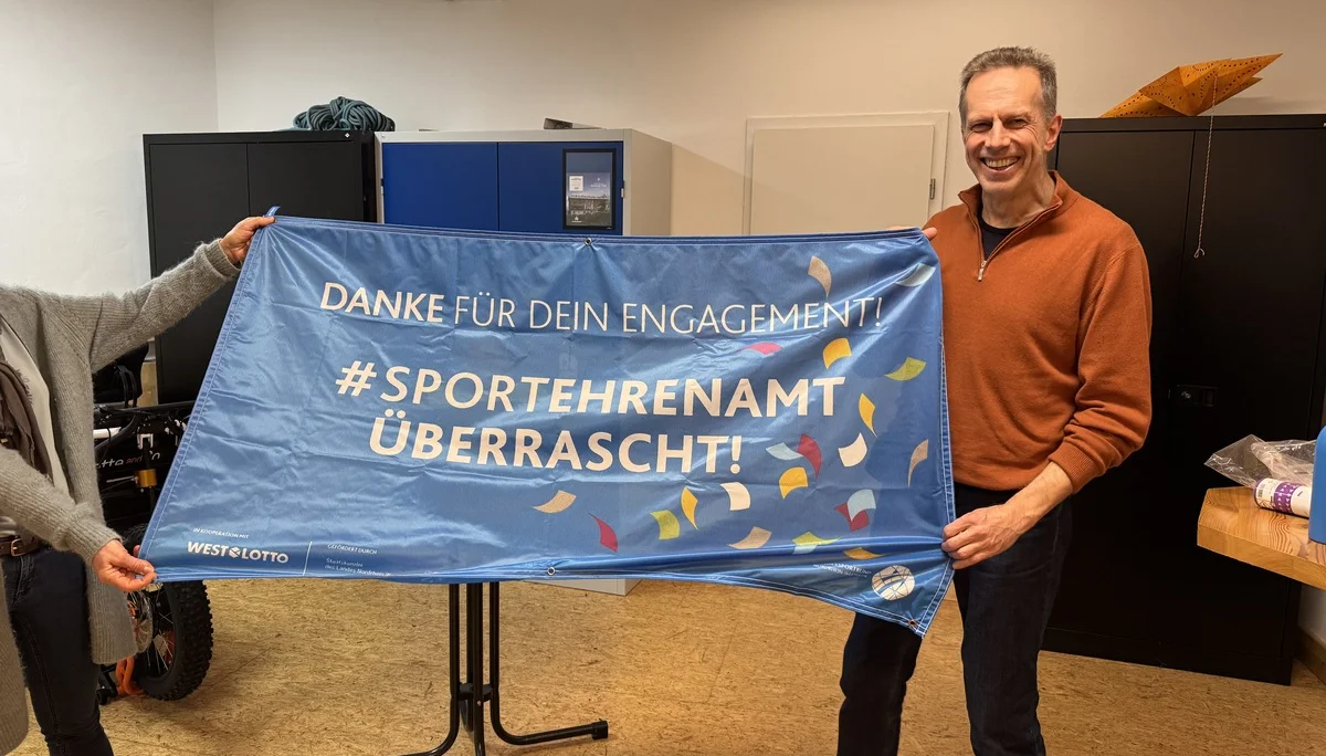 Mann hält ein Banner mit der Aufschrift „#Sportehramt überrascht“ in einem Raum mit anderen Anwesenden. | © DAV Dortmund