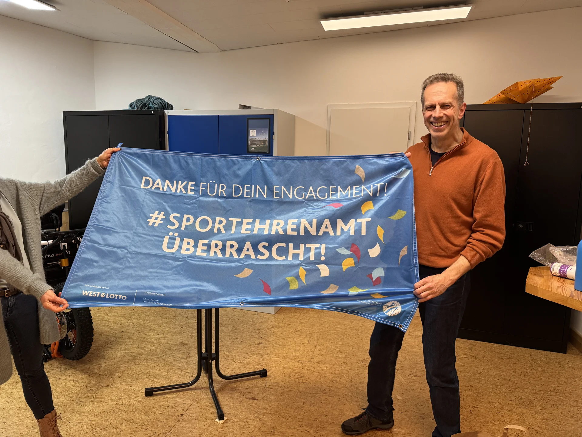 Mann hält ein Banner mit der Aufschrift „#Sportehramt überrascht“ in einem Raum mit anderen Anwesenden. | © DAV Dortmund