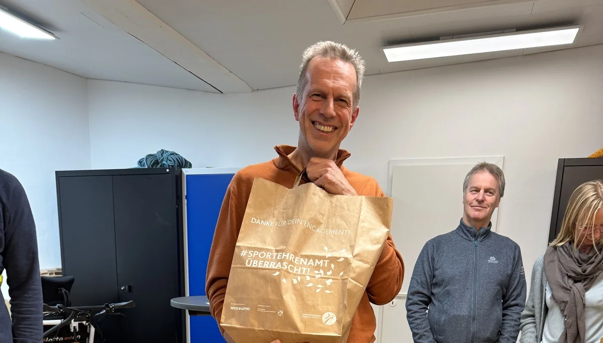 Mann hält eine Papiertüte mit Geschenk, umringt von lachenden Personen in einem Raum. | © DAV Dortmund