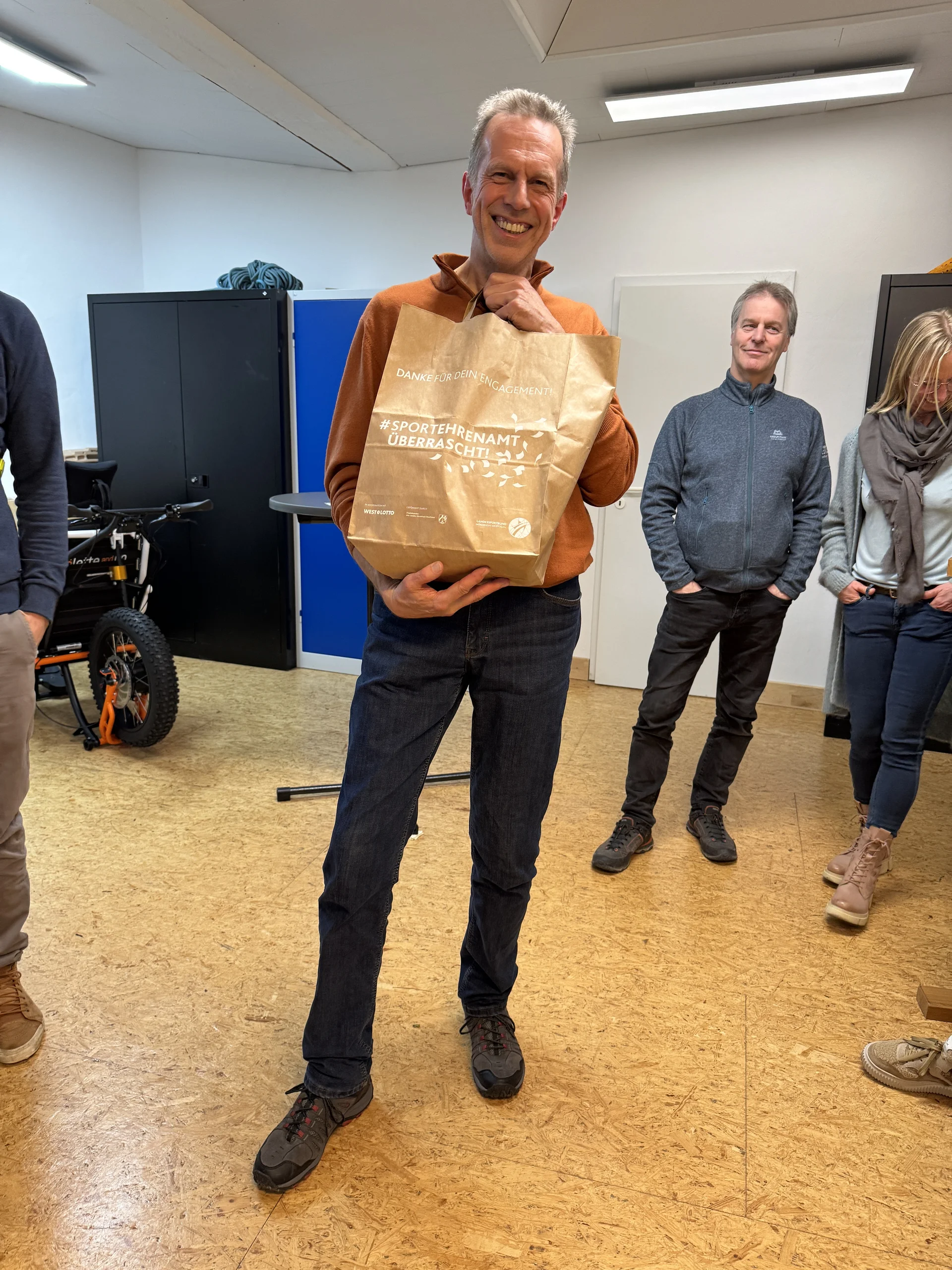 Mann hält eine Papiertüte mit Geschenk, umringt von lachenden Personen in einem Raum. | © DAV Dortmund