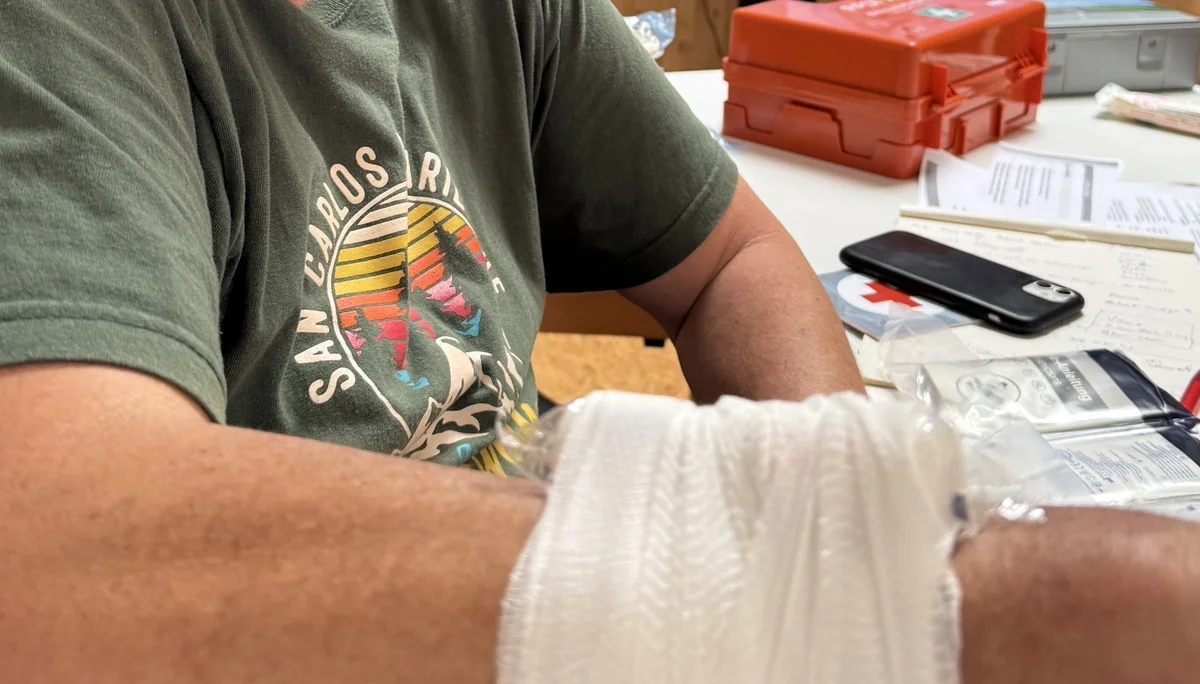 Ein Mann zeigt seinen Arm mit einem frisch angelegten Verband, im Hintergrund läuft eine Erste-Hilfe-Präsentation. | © DAV Dortmund