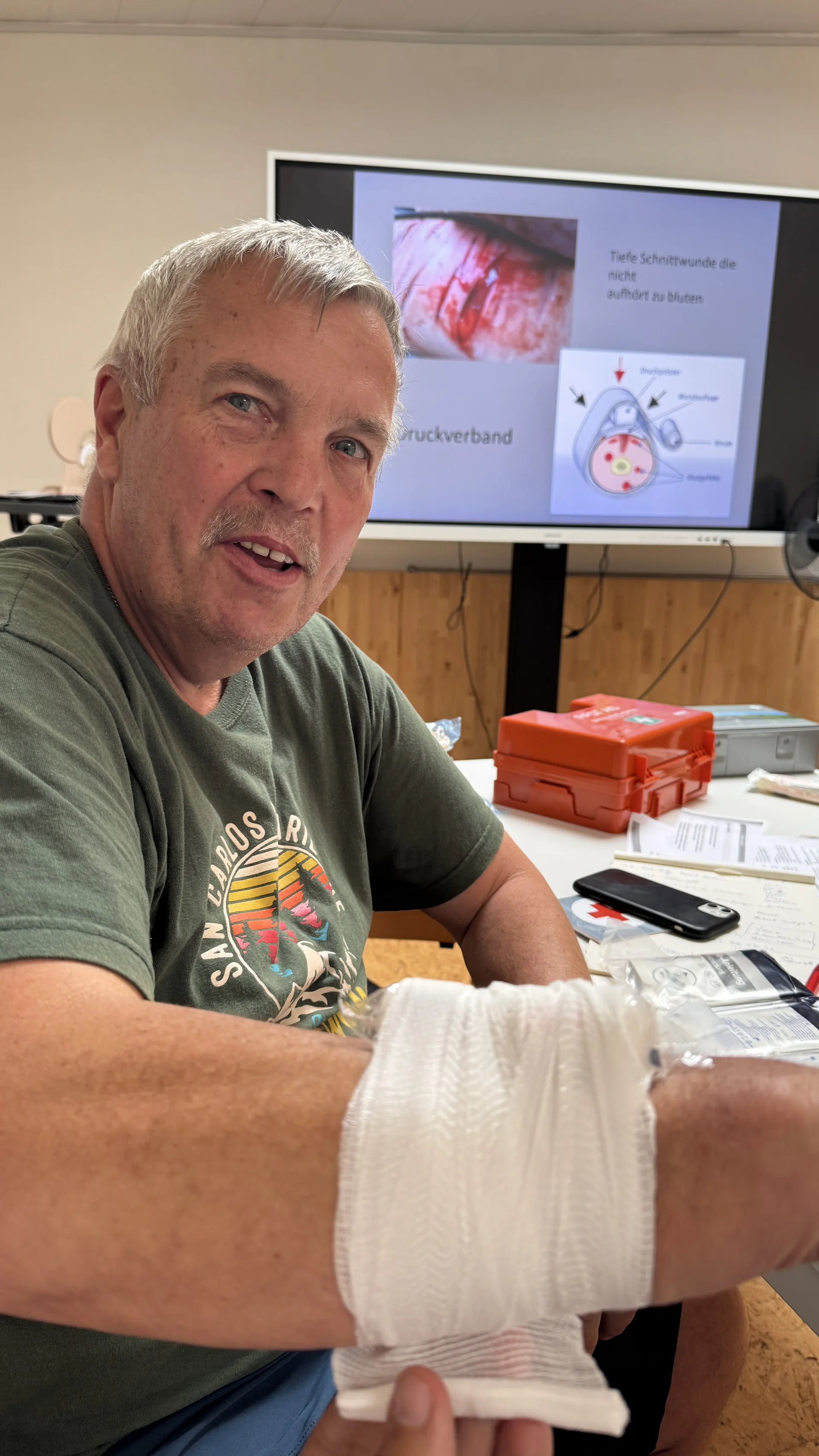 Ein Mann zeigt seinen Arm mit einem frisch angelegten Verband, im Hintergrund läuft eine Erste-Hilfe-Präsentation. | © DAV Dortmund