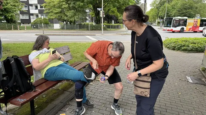 Eine Frau sitzt auf einer Bank, während ein Mann ihren Knieschutz überprüft und eine andere Frau daneben steht. | © DAV Dortmund