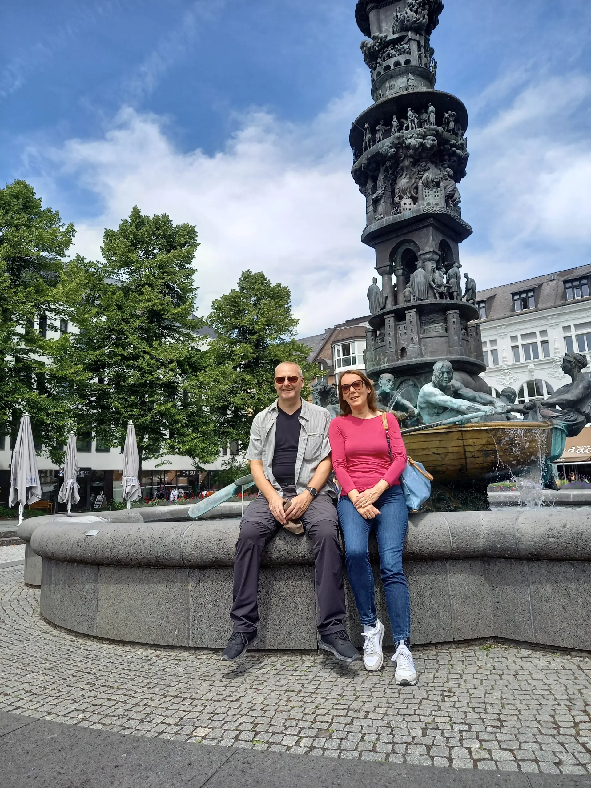Zwei Personen sitzen auf dem Rand eines Brunnens mit kunstvoller Statue auf einem Stadtplatz. | © DAV Dortmund