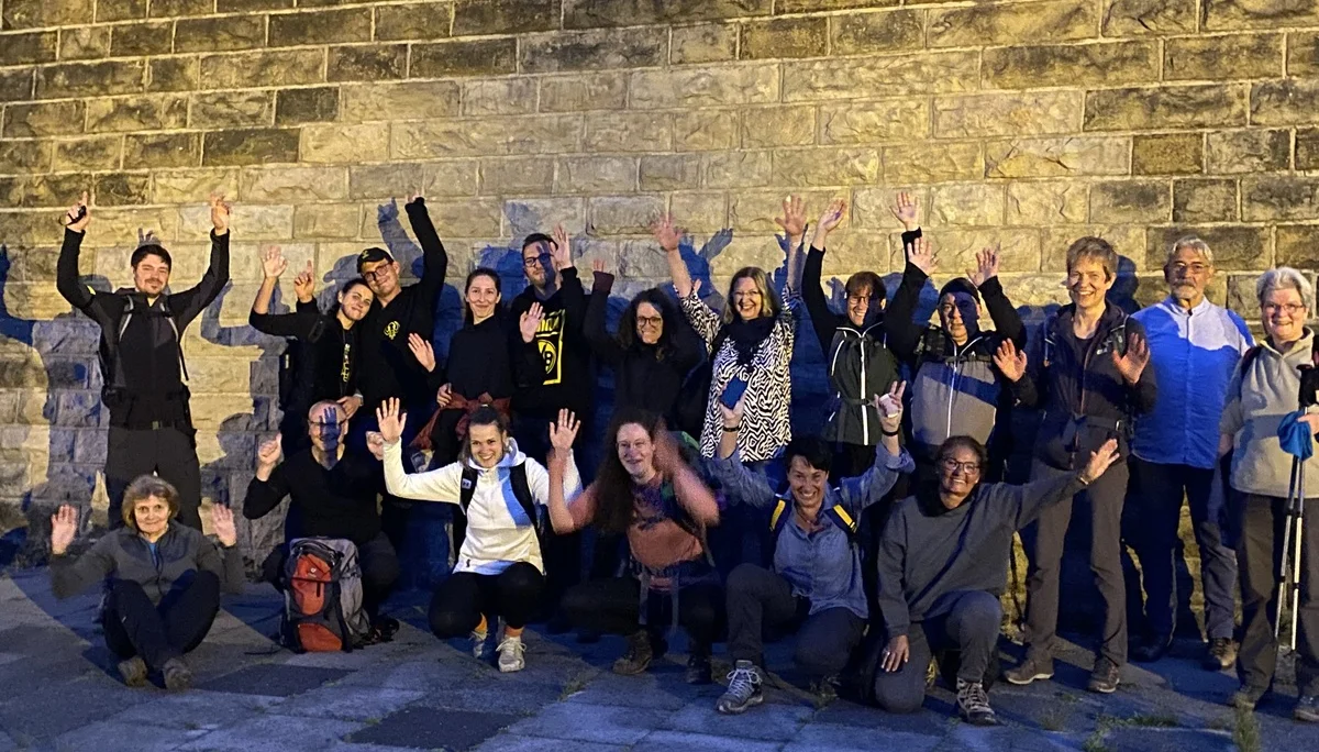 Eine große Wandergruppe posiert lachend und mit erhobenen Armen vor einer Steinmauer bei Nacht. | © DAV Dortmund