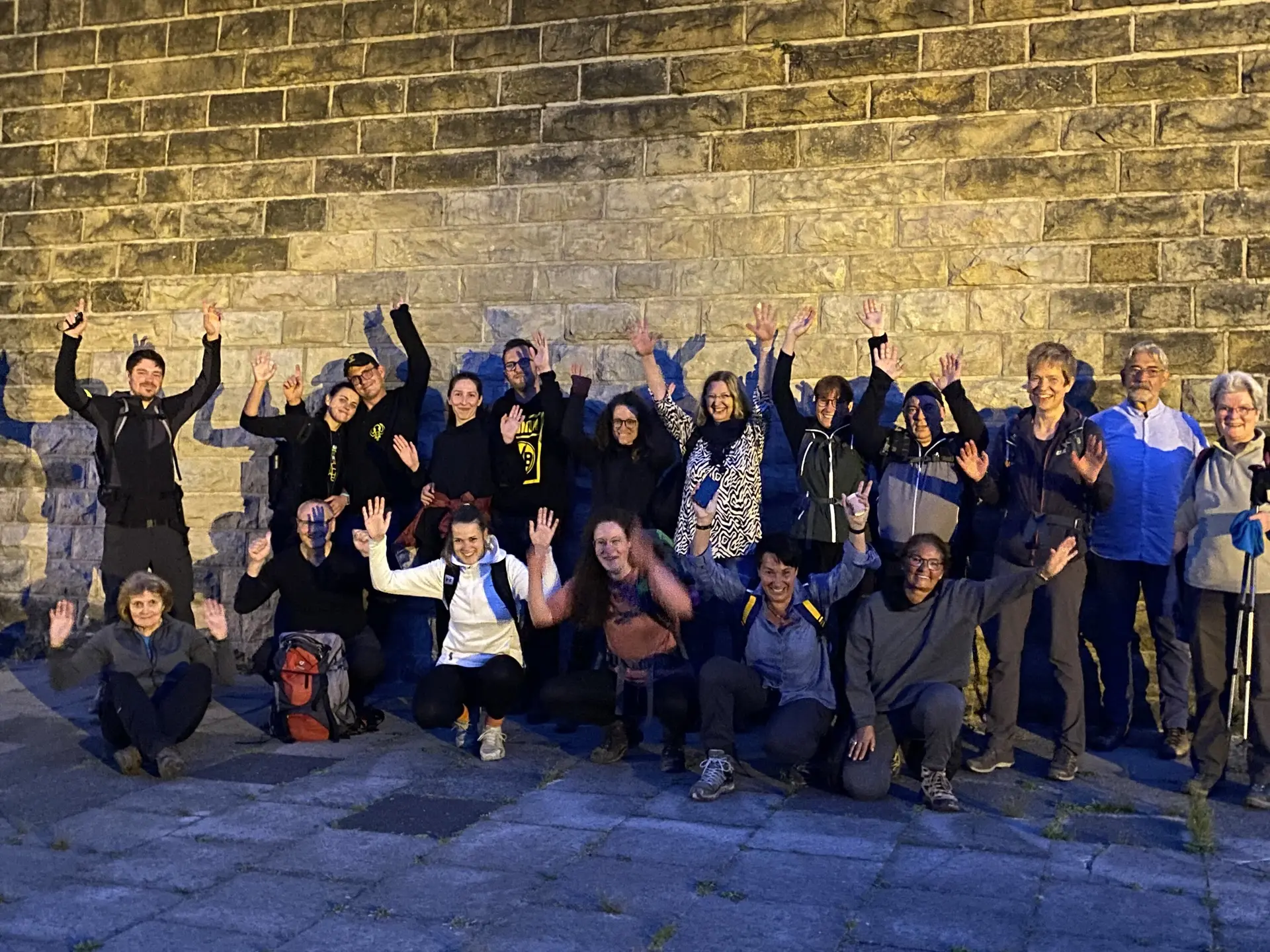Eine große Wandergruppe posiert lachend und mit erhobenen Armen vor einer Steinmauer bei Nacht. | © DAV Dortmund