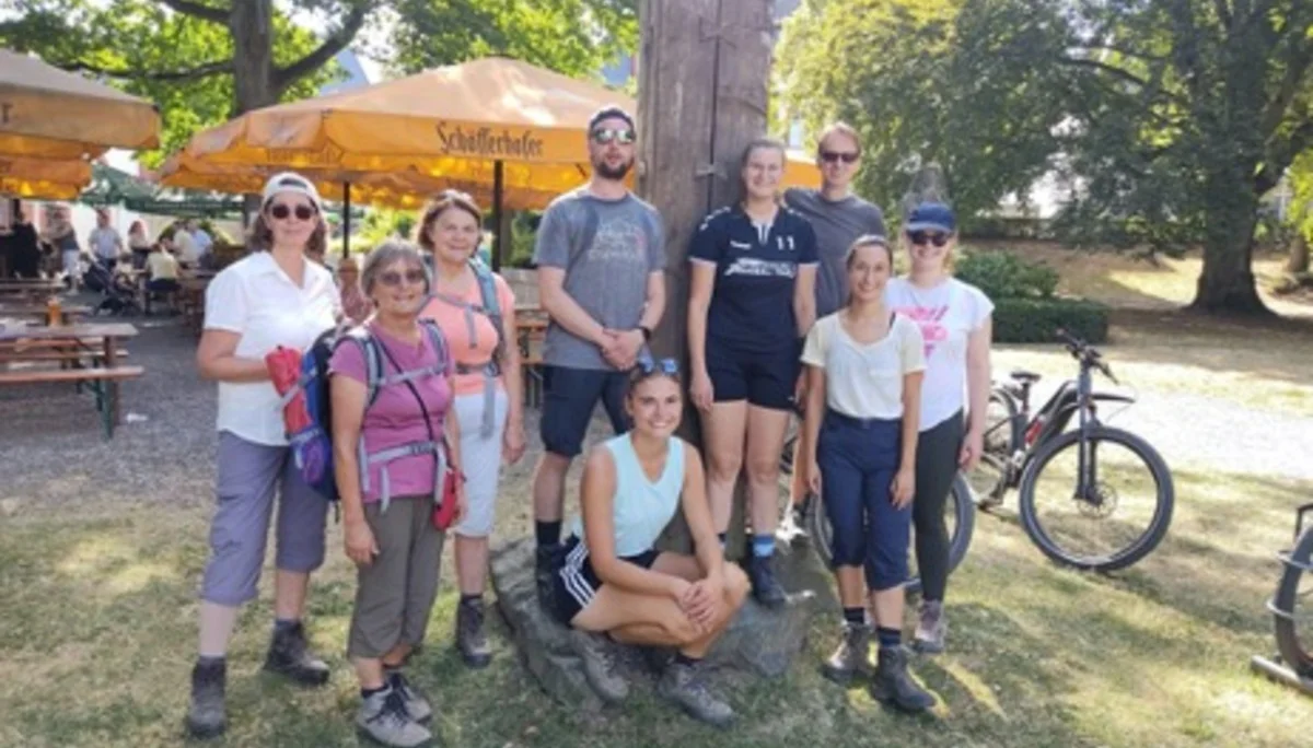 Eine Gruppe von Wandernden steht bei einem Holzpfosten auf einer Wiese mit Biergarten im Hintergrund. | © DAV Dortmund