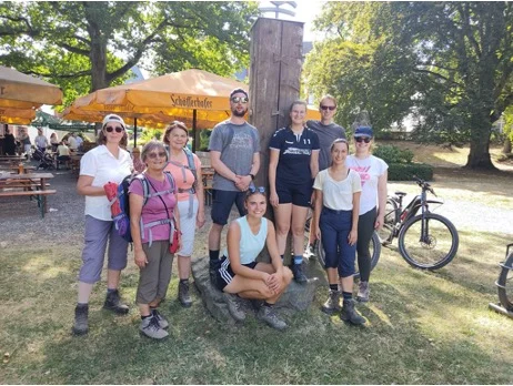 Eine Gruppe von Wandernden steht bei einem Holzpfosten auf einer Wiese mit Biergarten im Hintergrund. | © DAV Dortmund