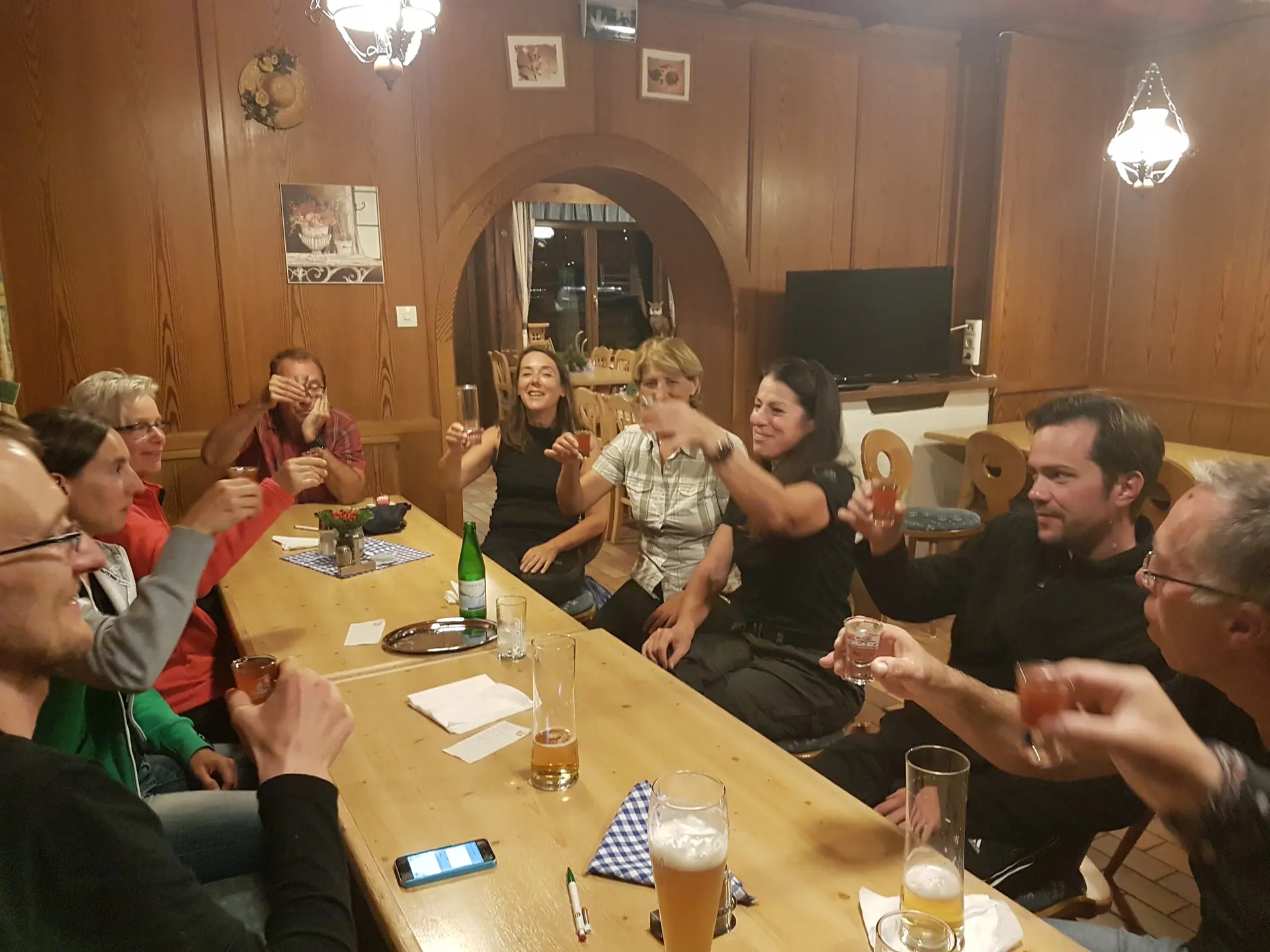 Gruppe sitzt gemeinsam am Holztisch in einer Hütte, lacht und stößt mit Getränken an. | © DAV Dortmund