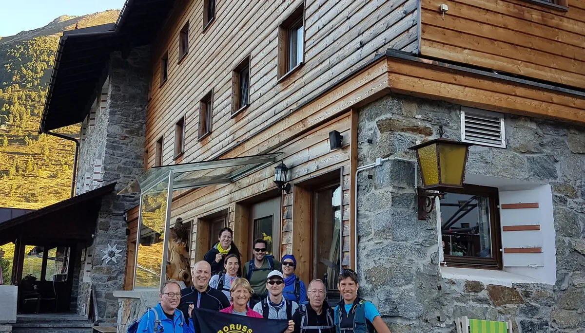 Gruppe steht vor einer großen Berghütte mit Holzfassade, einige halten eine Vereinsfahne. | © DAV Dortmund