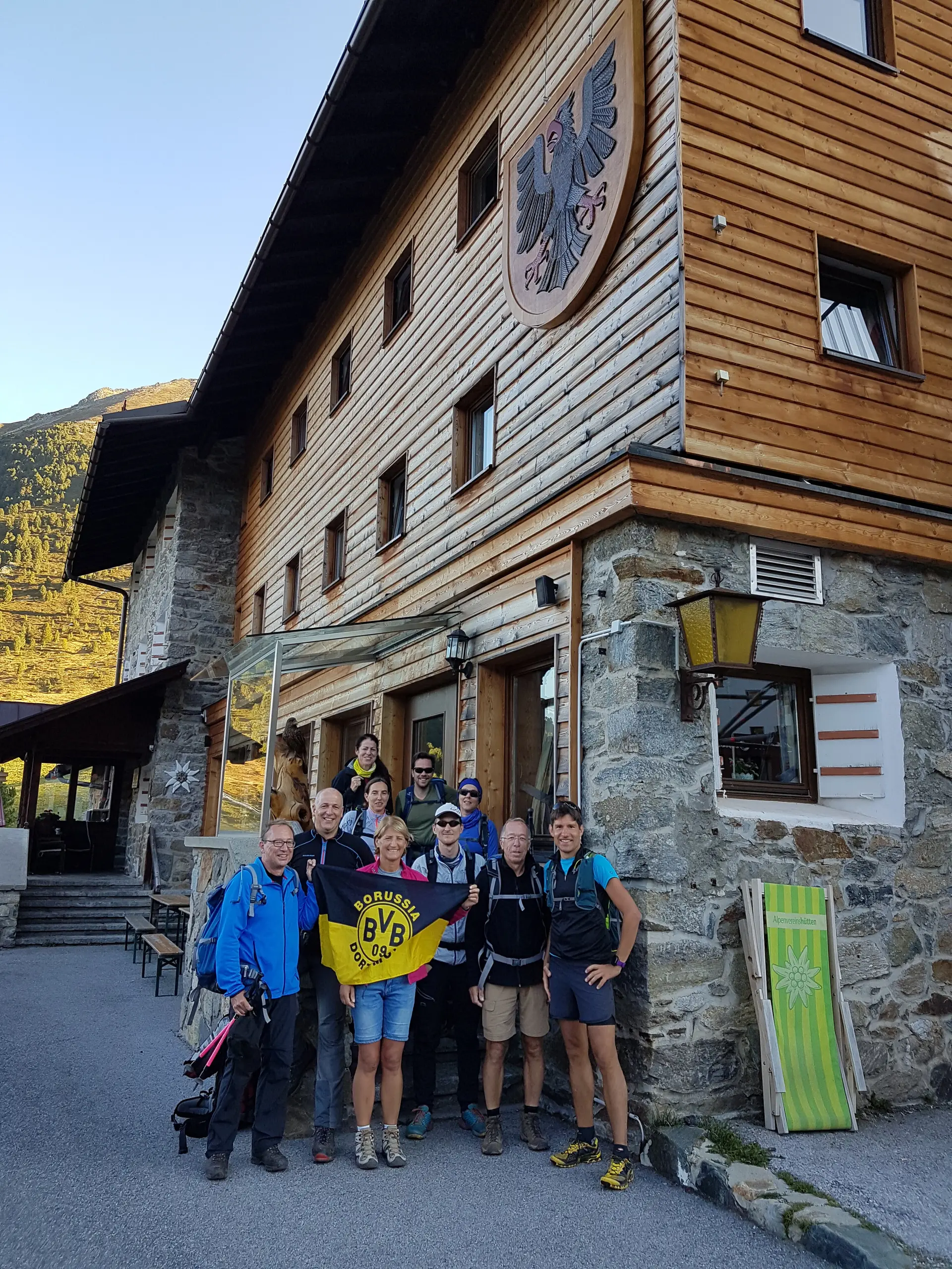 Gruppe steht vor einer großen Berghütte mit Holzfassade, einige halten eine Vereinsfahne. | © DAV Dortmund