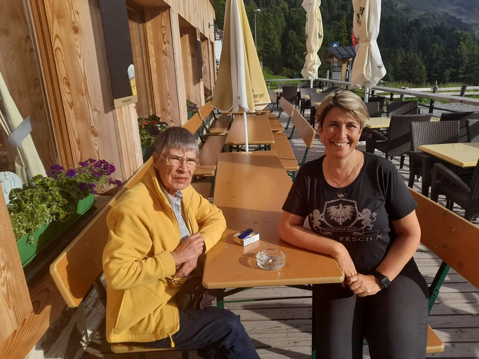Zwei Personen sitzen an einem Holztisch auf der Sonnenterrasse einer Berghütte bei warmem Wetter. | © DAV Dortmund