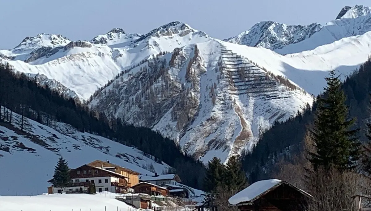 Mehrere Holzhütten liegen am Hang eines sonnigen Tals mit schneebedeckten Bergrücken im Hintergrund. | © DAV Dortmund