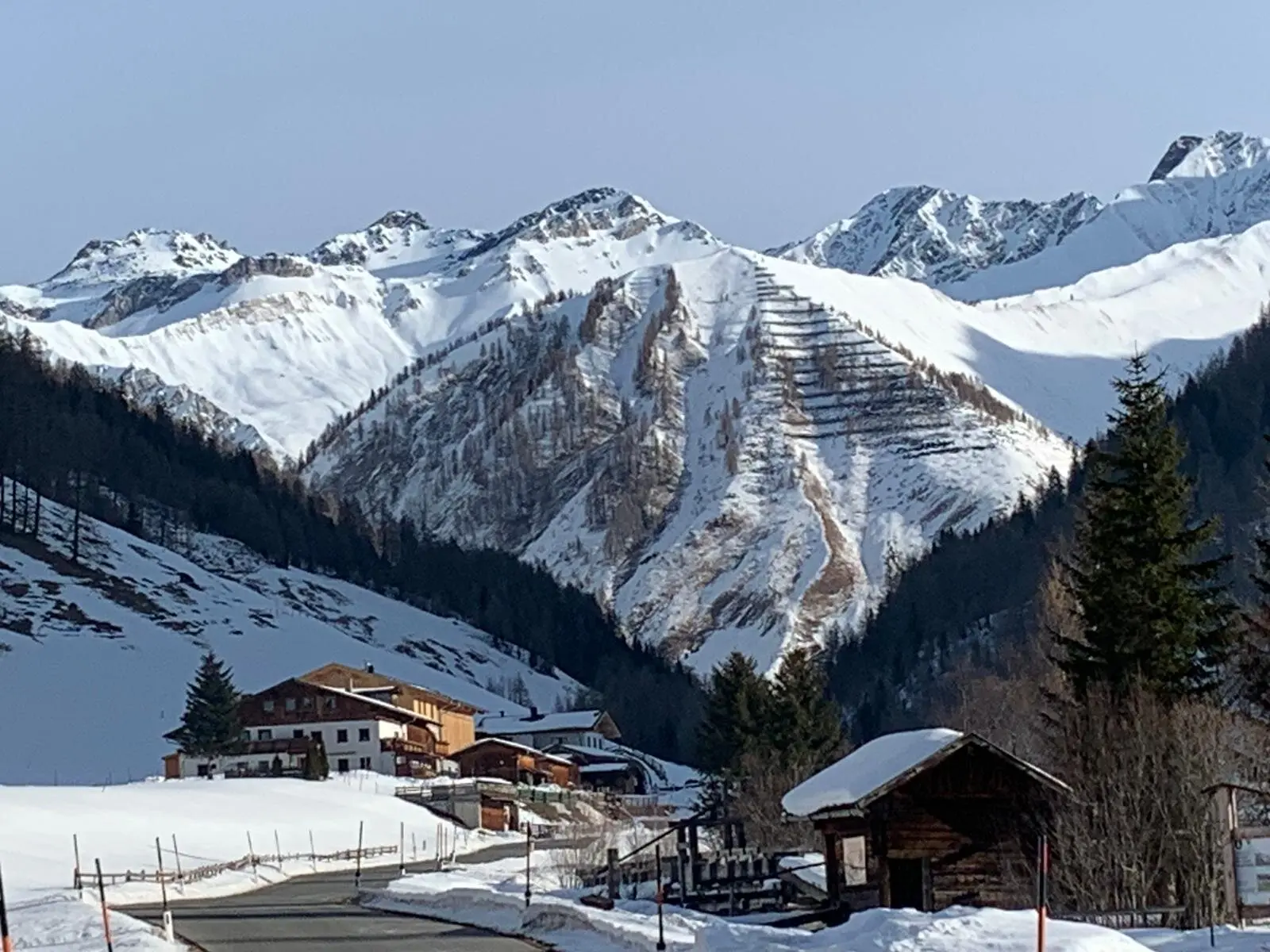 Mehrere Holzhütten liegen am Hang eines sonnigen Tals mit schneebedeckten Bergrücken im Hintergrund. | © DAV Dortmund