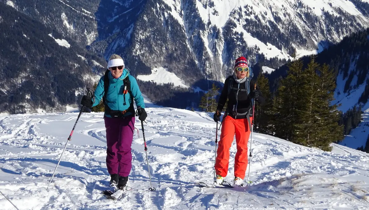 Zwei Frauen mit Tourenski gehen lachend einen sonnigen Hang mit verschneiten Bergen im Hintergrund hinauf. | © DAV Dortmund