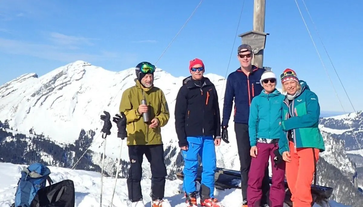 Fünf Personen stehen bei Sonnenschein mit Tourenski am Gipfelkreuz, Rucksäcke und Skier liegen im Schnee. | © DAV Dortmund