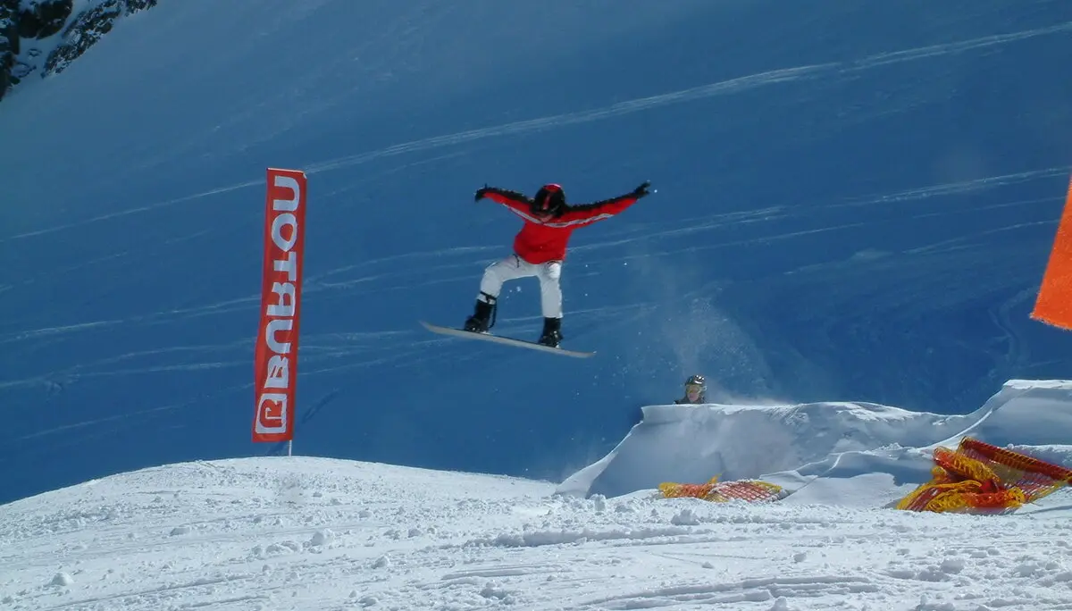 Eine Person springt mit einem Snowboard über eine Rampe im Schneepark, im Hintergrund ein Banner mit „Burton“. | © DAV Dortmund
