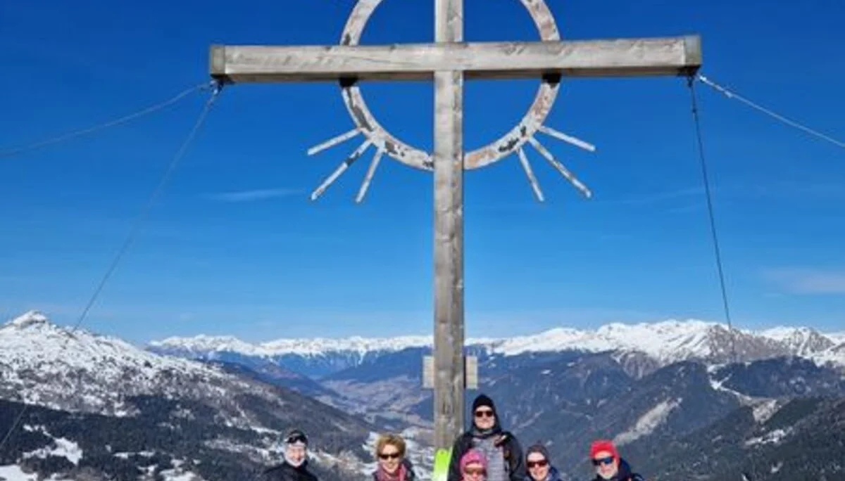 Eine Skitourengruppe steht unter einem Gipfelkreuz mit Strahlen in einer verschneiten Alpenlandschaft. | © DAV Dortmund