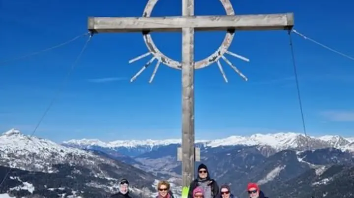 Eine Skitourengruppe steht unter einem Gipfelkreuz mit Strahlen in einer verschneiten Alpenlandschaft. | © DAV Dortmund