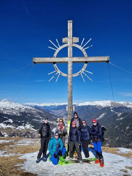 Eine Skitourengruppe steht unter einem Gipfelkreuz mit Strahlen in einer verschneiten Alpenlandschaft. | © DAV Dortmund