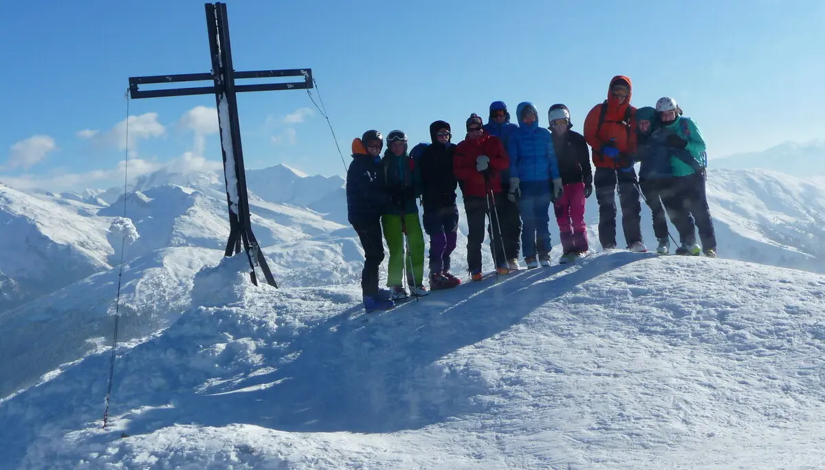 Jugendliche in bunter Winterkleidung stehen auf einem verschneiten Gipfel mit schwarzem Gipfelkreuz. | © DAV Dortmund