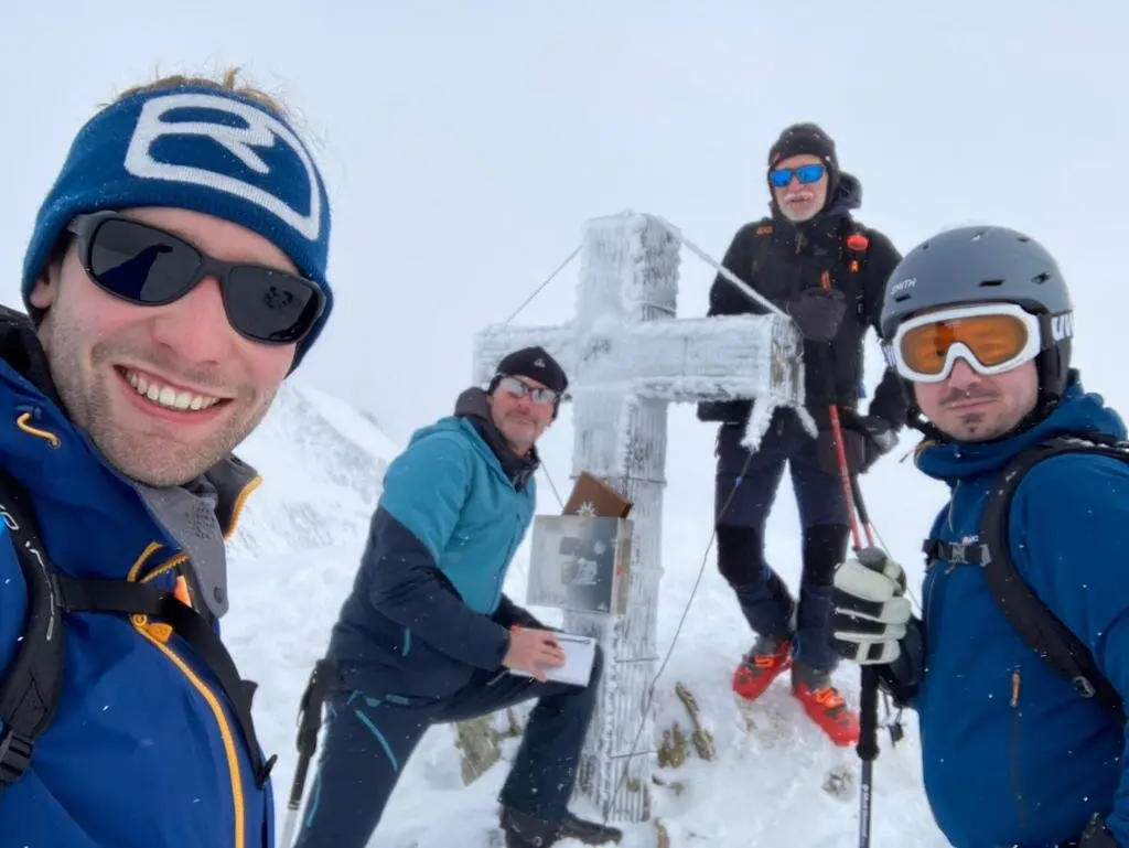 Vier Skibergsteiger posieren an einem mit Eis bedeckten Gipfelkreuz bei Schneefall. | © DAV Dortmund