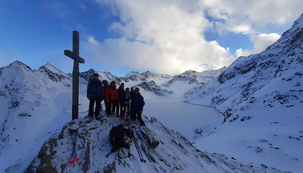 Gruppe steht am Gipfelkreuz mit Blick auf einen gefrorenen See inmitten schneebedeckter Berge. | © DAV Dortmund