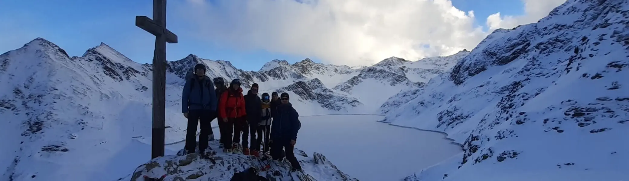 Gruppe steht am Gipfelkreuz mit Blick auf einen gefrorenen See inmitten schneebedeckter Berge. | © DAV Dortmund