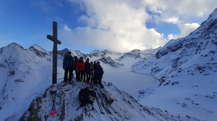 Gruppe steht am Gipfelkreuz mit Blick auf einen gefrorenen See inmitten schneebedeckter Berge. | © DAV Dortmund