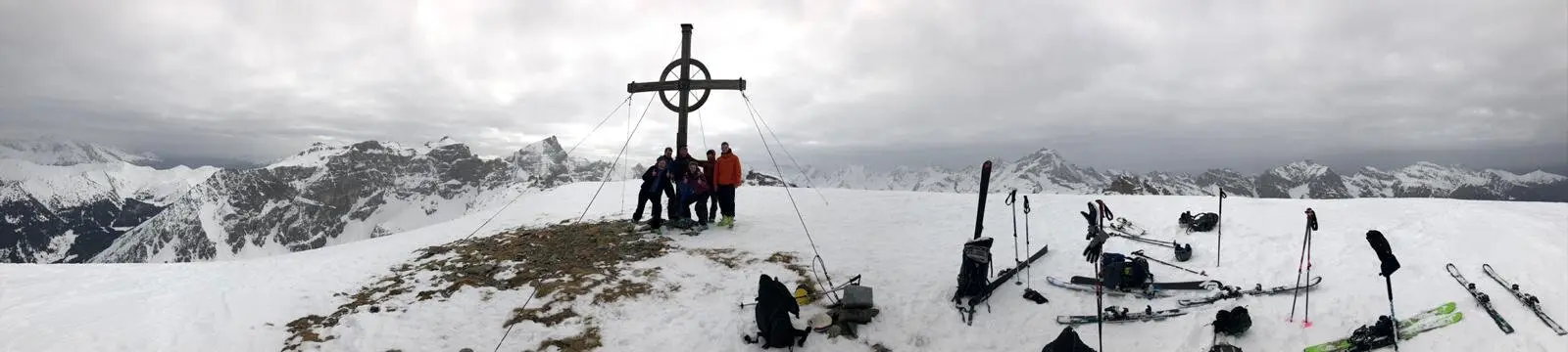 Breites Panorama einer Skitourengruppe am Gipfelkreuz mit abgelegten Skiern und verschneiter Bergkulisse. | © DAV Dortmund