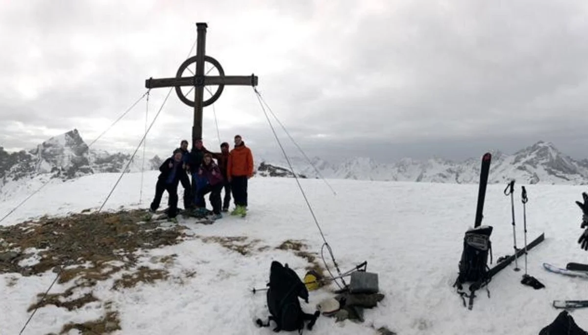 Fünf Skitourengeher*innen posieren am Gipfelkreuz mit Blick auf schneebedeckte Berge. | © DAV Dortmund
