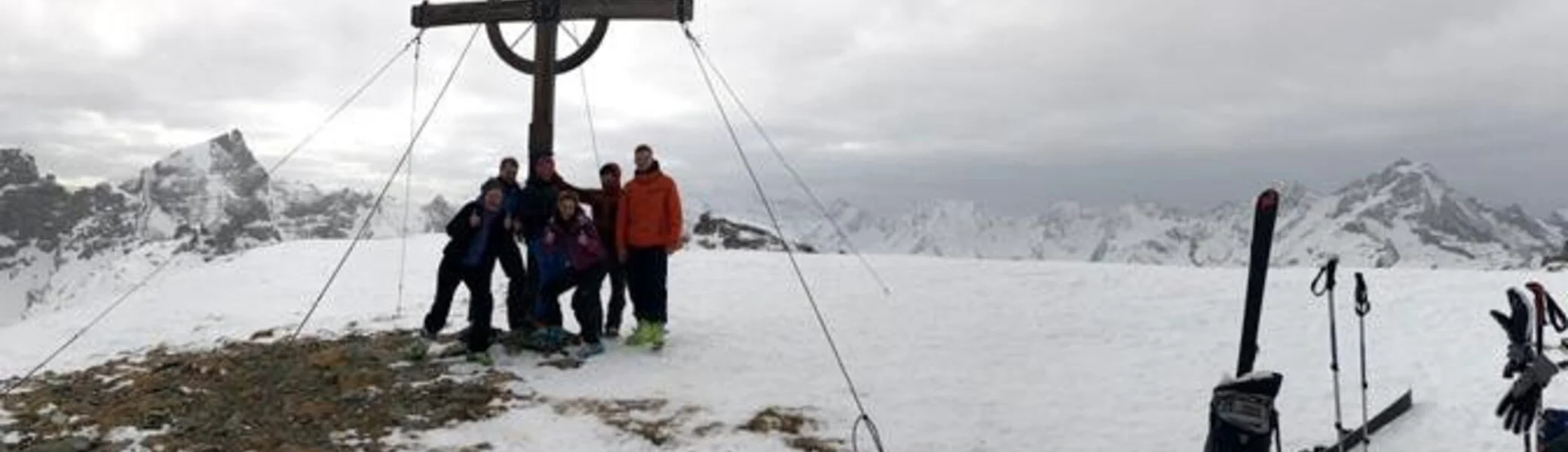 Fünf Skitourengeher*innen posieren am Gipfelkreuz mit Blick auf schneebedeckte Berge. | © DAV Dortmund